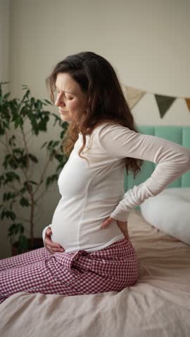 Pregnant woman in her third trimester suffering from lower back pain, sitting on a bed and massaging her lumbar region while caressing her belly to soothe the discomfort of expecting a baby