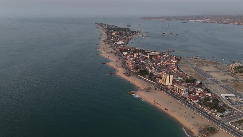 high-angle aerial perspective of the Ilha do Cabo peninsula in Luanda, Angola, showing the long sandy beach on the Atlantic side and the ship-filled bay on the other