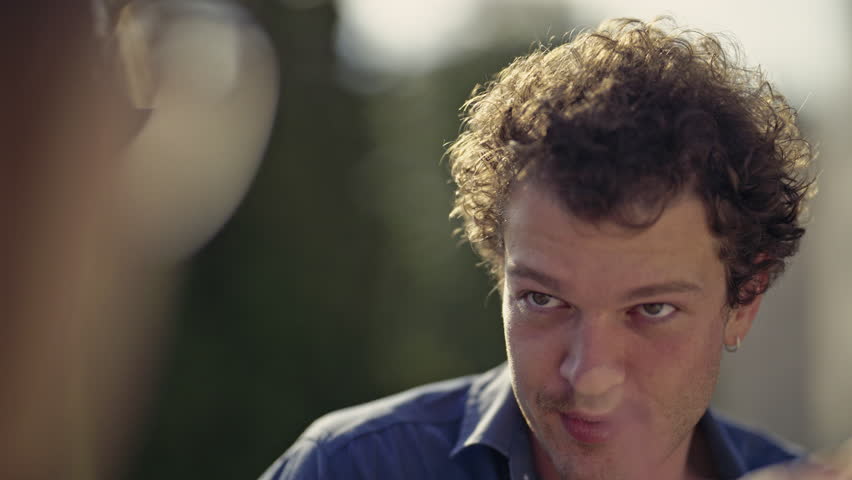 Captivating close-up of an expressive young man with curly hair, animatedly conversing and gesturing outdoors. His joyful expressions suggest a friendly chat or engaging storytelling.
