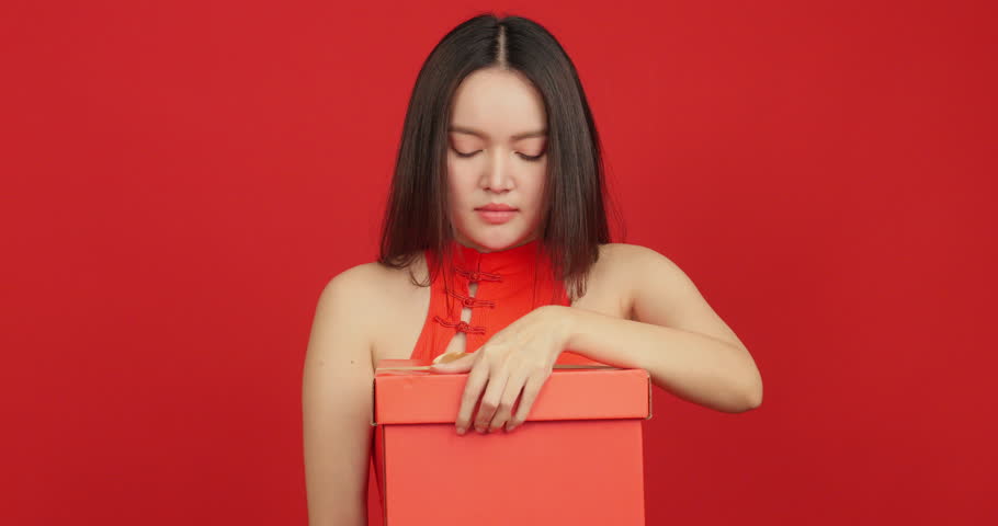 Happy young Asian woman in red Chinese dress opening a gift box on red background.