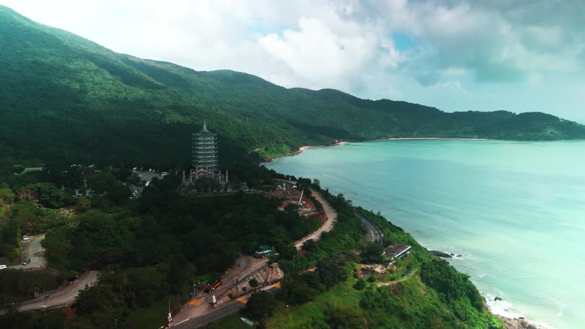 Da nang aerial view of linh ung pagoda's tiered tower on lush son tra peninsula beside winding coastal road, blending buddhist landmark with tranquil blue ocean landscape