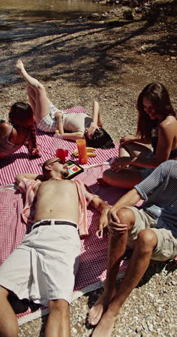 A group of diverse young friends unwind and share joyful moments during a sunny picnic by the water. They are enjoying the warm weather and each other