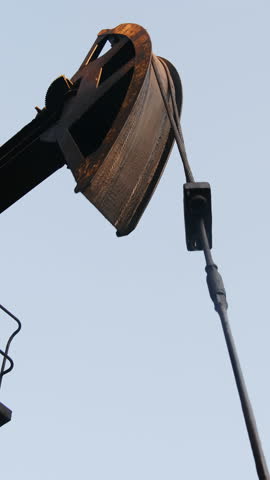 Horse head of old pumpjack moves up and down at oilfield against blue sky vertical shot. Vintage equipment to pump crude oil from well closeup
