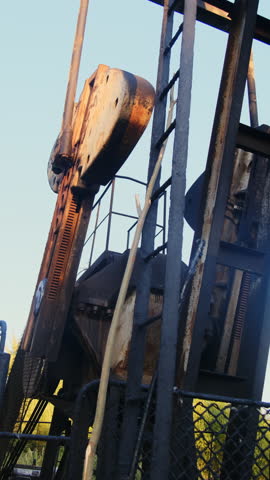 Derrick pumps crude oil from pit under blue sky slow motion. Old nodding donkey extracts fossil fuel from onshore well at countryside vertical shot