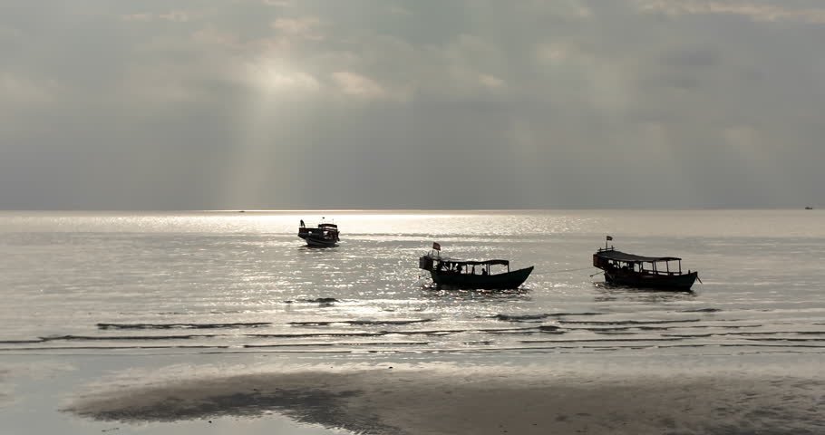 Silhouetted Cambodian boats on a shimmering sea under dramatic sunbeams at sunset.
