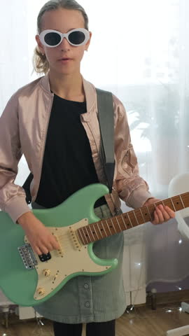 Vertical video. Teen girl in her room wearing sunglasses plays an electric guitar, having fun with her music and focusing on her creative hobby, slow motion