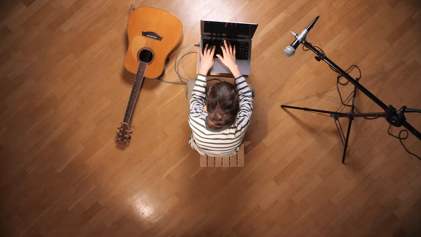 Boy sitting on a stool, typing on a laptop with an acoustic guitar and a microphone on a stand arranged on the parquet floor, representing creativity and digital music production