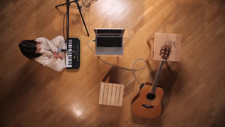 Woman playing a digital keyboard and singing into a microphone, connected to a laptop on a wooden floor with an acoustic guitar illustrating the process of creating music