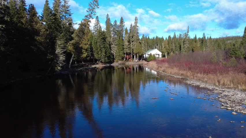 A peaceful river meanders past a white cottage on the edge of a coniferous forest. The calm water reflects the blue sky, while reeds line the bank, offering a haven of rural tranquility. Quebec, Canada, 2025.
