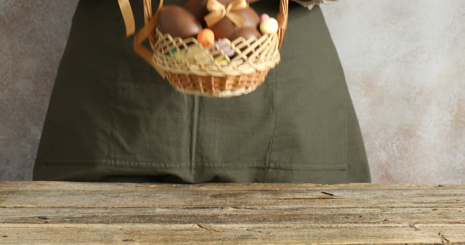 Woman holding wicker basket with chocolate Easter bunny and eggs at wooden table on light grey background, closeup