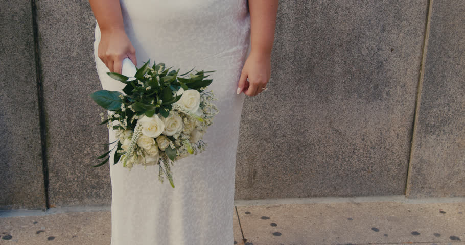 A beautiful bride in a white wedding dress walks forward and looks into the camera with a smile against the background of a stone building.