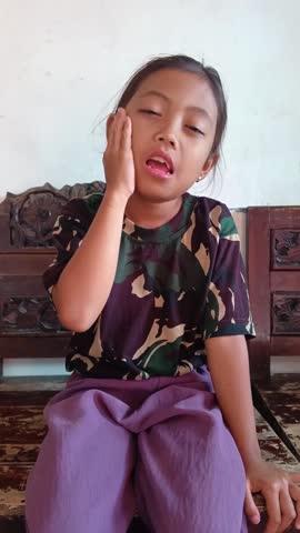 A young Asian girl expressing pain while holding her cheek due to a toothache, sitting on a wooden bench in a domestic setting.