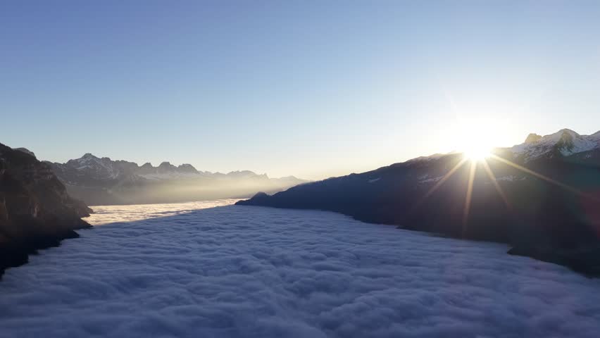Aerial sunrise over a valley filled with clouds in the Swiss Alps, with bright sun starburst above snowy mountain ridge.