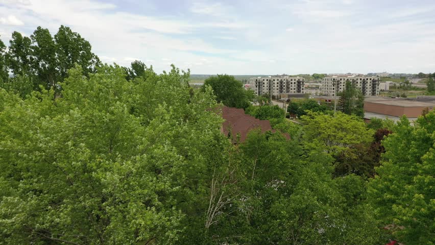 Ascending drone footage revealing a sprawling suburban town with residential housing, apartment buildings, roads with traffic, and a distant lake under a partly cloudy summer sky. Barrie ontario
