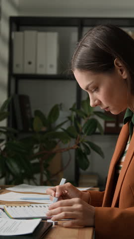 Vertical shot of Caucasian female office employee highlighting handwritten notes in black notebook at workplace desk, closing it and putting back in drawer after finishing focused work or study