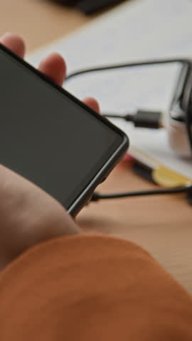 Vertical closeup of employee plugging smartphone into portable charger while working at wooden office desk surrounded by documents