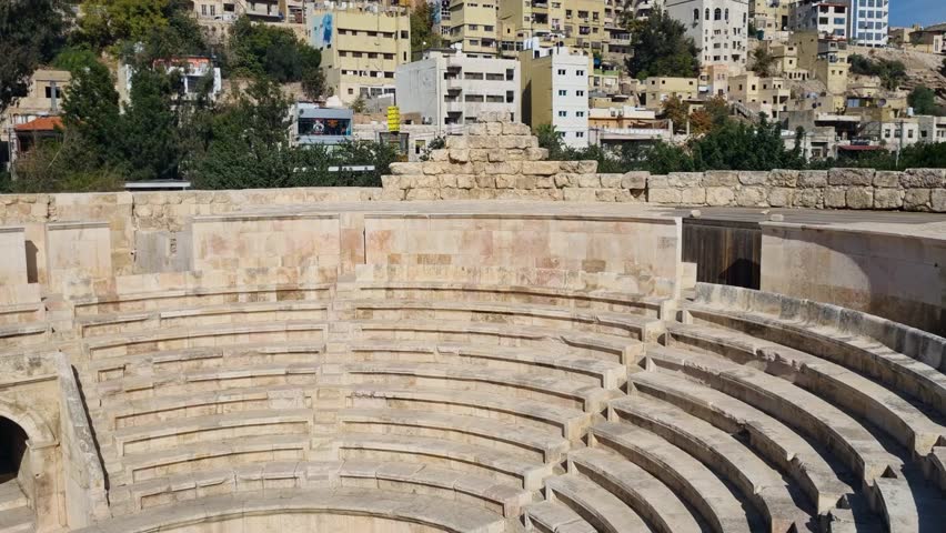 Clear daylight view inside the Roman Odeon Theater in Amman, Jordan, featuring ancient stone seating and preserved historical architecture, set against a bright blue sky with residential buildings