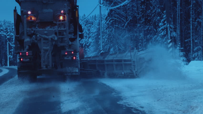 Clearing snow from the road in winter. Large industrial vehicle removes snow from the highway, sprinkles sand or chemicals. Polar night and snow storm, blizzard in arctic.