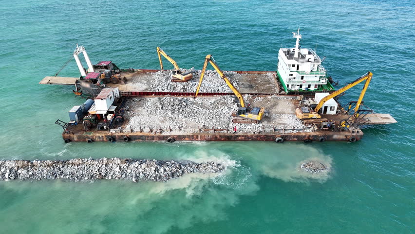 marine construction barge working at sea. Heavy machinery excavators unloading rocks to build a stone breakwater or sea wall foundation. Coastal civil engineering infrastructure project