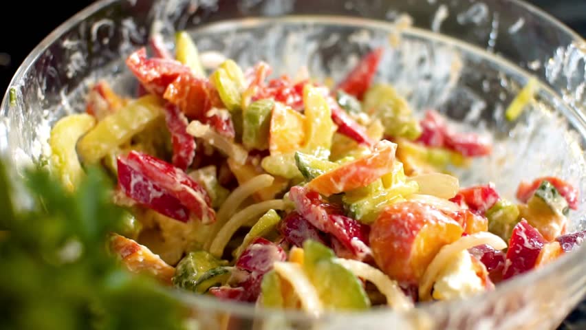 Close up shot of person mixing fresh vegetable salad with creamy white dressing in a glass bowl using two spoons in a bright kitchen