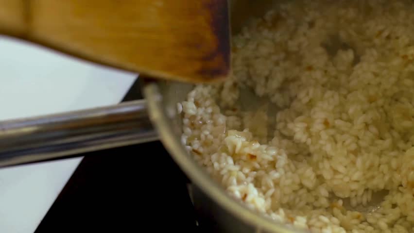 Liquid broth being poured into a stainless steel pot of rice and stirred with a wooden spoon to cook a creamy Italian risotto