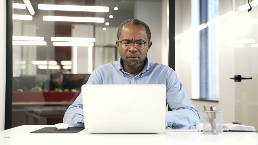 Upset worried african american businessman reads bad news on laptop sitting at desk at workplace in business office. Frustrated worker is sad reviewing negative information on a computer. Front view