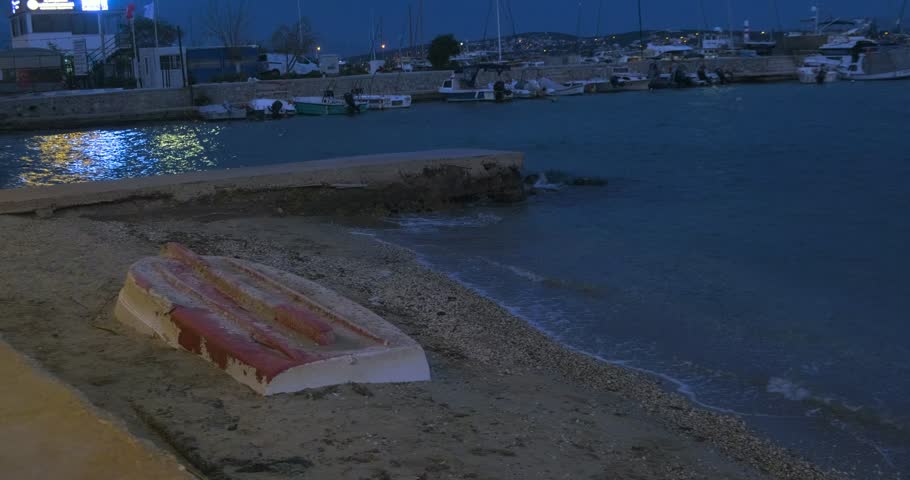 A small, weathered dinghy or rowboat pulled up and overturned on the sand. Nighttime scene with a concrete pier and calm blue water in the background. Harbor lights reflect on the water. Fishing village concept.