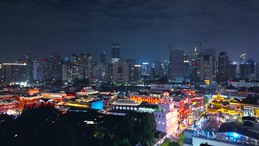 Aerial shot of illuminated traditional architecture and rooftops within Yu Garden, contrasting with surrounding modern high-rise skyscrapers in Shanghai, China.
