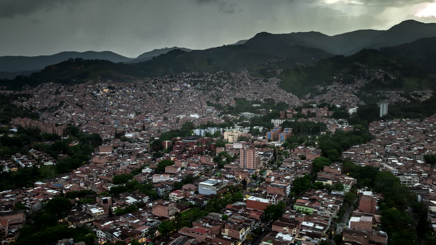 Drone hyperlapse over Medellín, Colombia at dusk, revealing dense hillside neighborhoods and city blocks framed by green mountains under stormy evening skies