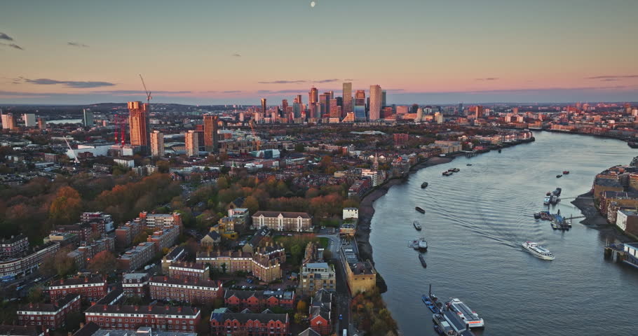 London cityscape panorama: Canary Wharf skyscrapers, residential buildings, and River Thames winding through urban areas with boats navigating at beautiful sunset. Aerial drone flight footage