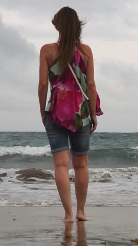 Woman from behind standing barefoot on the wet sand as waves roll in on a cloudy day