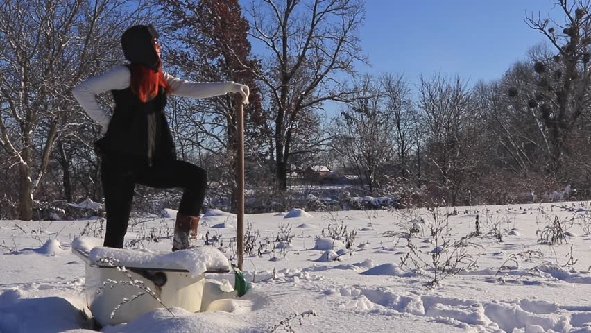 A woman in winter clothes stands in a snowy bathtub in the yard, leaning on a shovel, looking into the distance on a frosty sunny day.