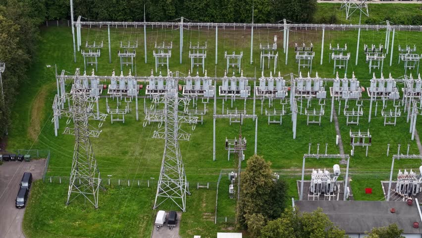 Aerial view of a high-voltage electrical substation with transformers, switchgear and steel pylons on green grass, showing modern electricity infrastructure and grid operations.