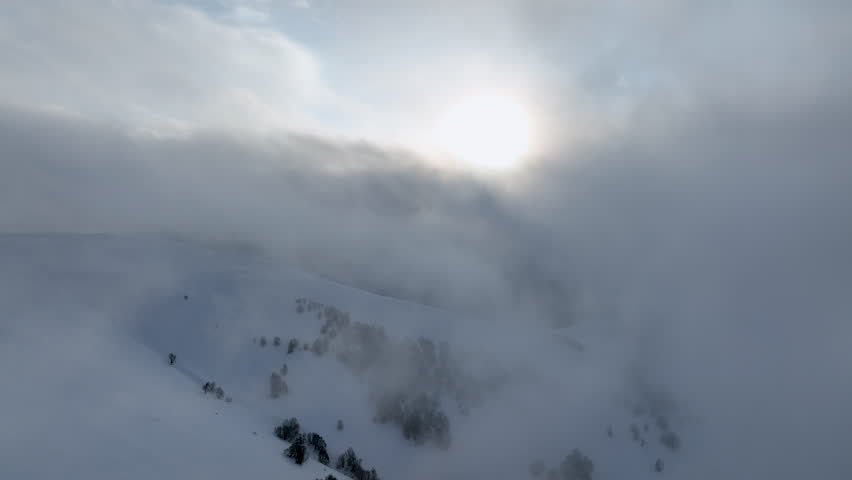 A dramatic aerial view of a winter landscape in Bakuriani resort featuring a bright sun piercing through thick clouds over snowy mountain peaks and a dark dense forest.