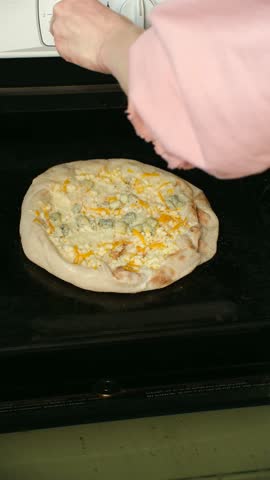 Woman wearing an oven mitt carefully places a delicious homemade pizza, topped with various cheeses, onto the rack of a preheated oven and closes the door to start the baking process. Vertical shot