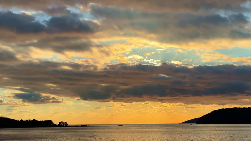 A serene sunset over calm water with gradually moving clouds and vibrant colors, a plane flying in the sky
