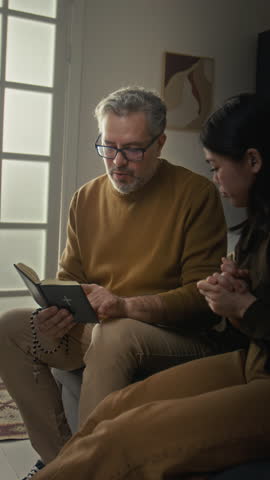 Vertical view of curious gen Z girl studying bible with middle-aged male religious teacher with glasses who visiting her at home