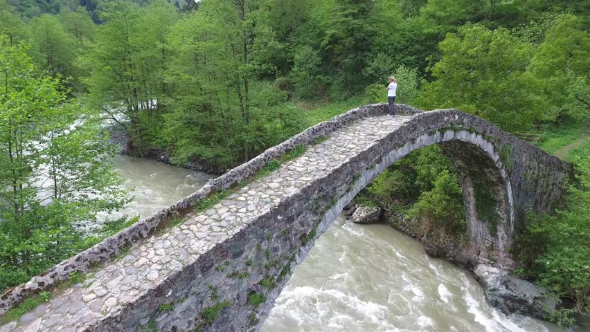 Aerial view shows blonde woman standing on historic stone arch bridge above forest river. Elevated perspective captures tourist enjoying scenic woodland and flowing water landscape.