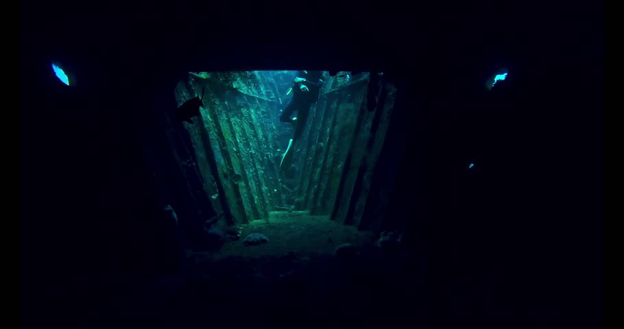 A lone scuba diver explores the dark interior of a sunken shipwreck, moving through rusty corridors in deep blue water. Cinematic slow motion underwater discovery.
