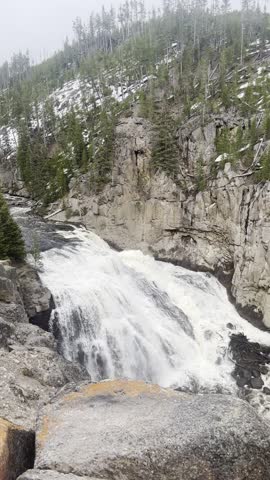 Water cascades over rocky cliffs along the Gibbon Falls area in Yellowstone National Park. A steady waterfall flows through a rugged canyon surrounded by evergreen forest, showcasing the natural landscape and flowing water of the park.