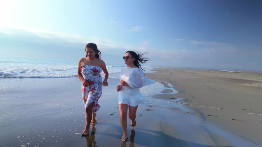 Two cheerful young female friends running barefoot on a sandy beach. Women enjoying their summer vacation together by the ocean shoreline