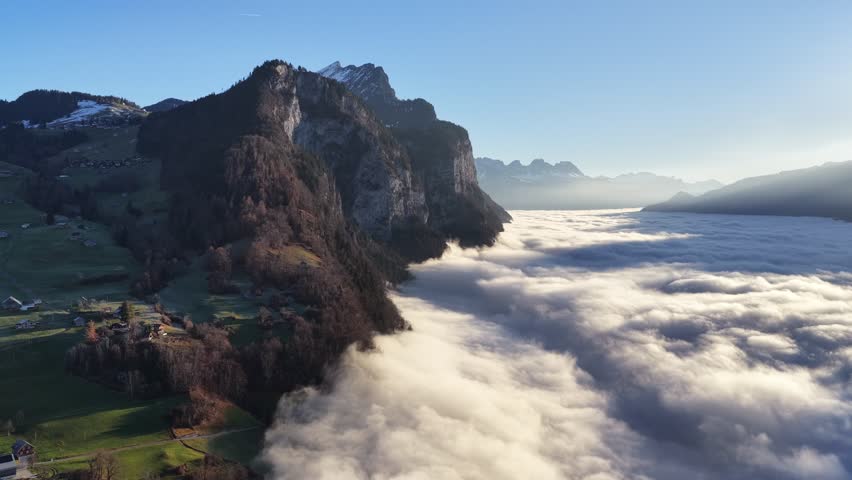 Aerial of Swiss houses on green slopes overlooking a vast sea of fog. Sharp mountain cliffs pierce the clouds under a bright sun in the Walensee region.