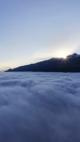 A breathtaking vertical aerial view of mountain cliffs piercing a thick sea of fog. Sunny alpine slopes and traditional houses overlook a vast cloud ocean in Switzerland.