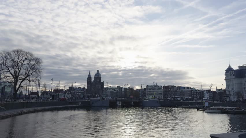 Wide waterfront shot near Amsterdam Centraal Station, showing the Basilica of Saint Nicholas, Prins Hendrikkade buildings, and reflections on the Open Havenfront under a bright cloudy sky