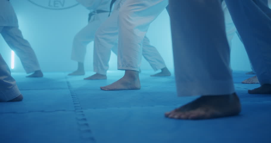 A group of students practices martial arts skills under soft blue lighting. They focus on their foot movements and stances. The trainers guide them during the session.