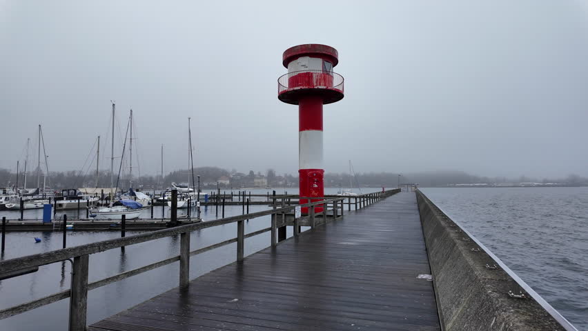 A scenic view of the red-and-white lighthouse at the harbor entrance in Eckernförde, Germany, captured on a misty, overcast day. A long wooden pier leads toward the lighthouse, with calm Baltic Sea