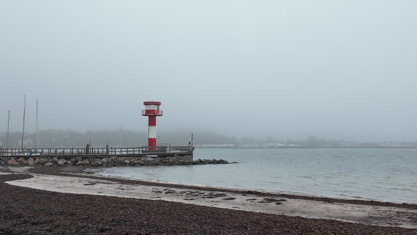 A wide coastal view featuring the red-and-white harbor lighthouse of Eckernförde, Germany, standing on a wooden pier along the Baltic Sea shoreline. The foreground shows a pebble beach and calm water