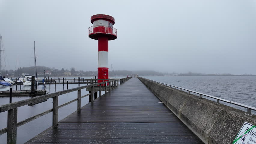 A scenic view of the red-and-white lighthouse at the harbor entrance in Eckernförde, Germany, captured on a misty, overcast day. A long wooden pier leads toward the lighthouse, with calm Baltic Sea