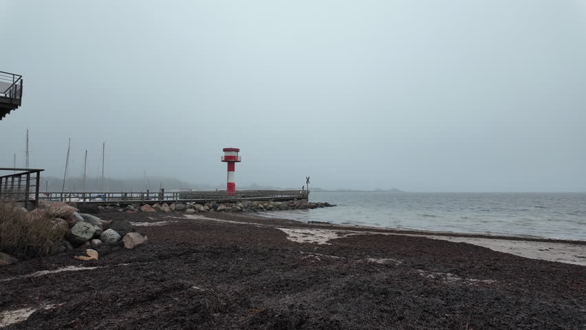 A wide coastal view featuring the red-and-white harbor lighthouse of Eckernförde, Germany, standing on a wooden pier along the Baltic Sea shoreline. The foreground shows a pebble beach and calm water