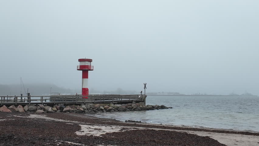 A wide coastal view featuring the red-and-white harbor lighthouse of Eckernförde, Germany, standing on a wooden pier along the Baltic Sea shoreline. The foreground shows a pebble beach and calm water